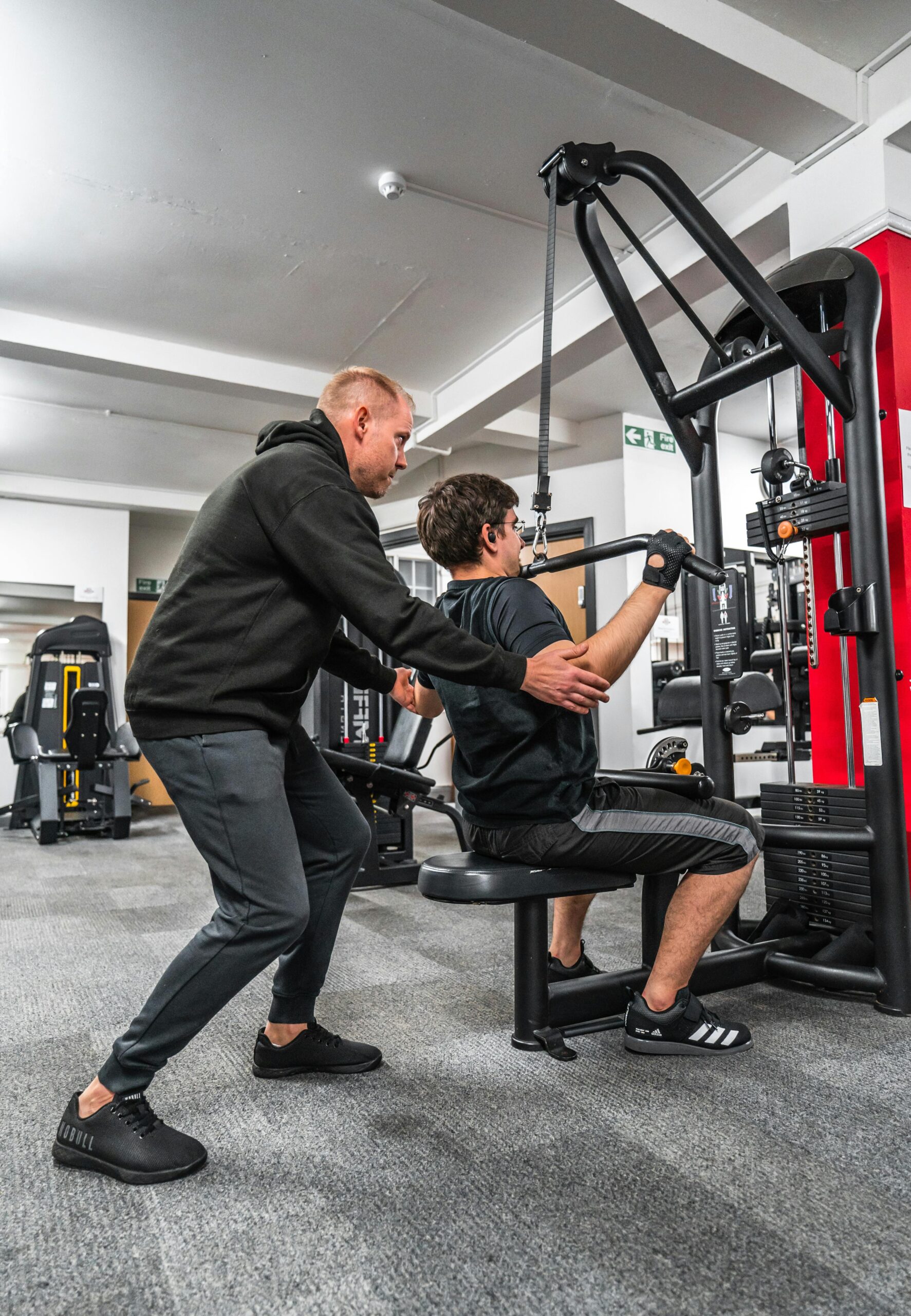 A personal trainer guides a client performing a workout using a gym machine, emphasizing correct form and technique.